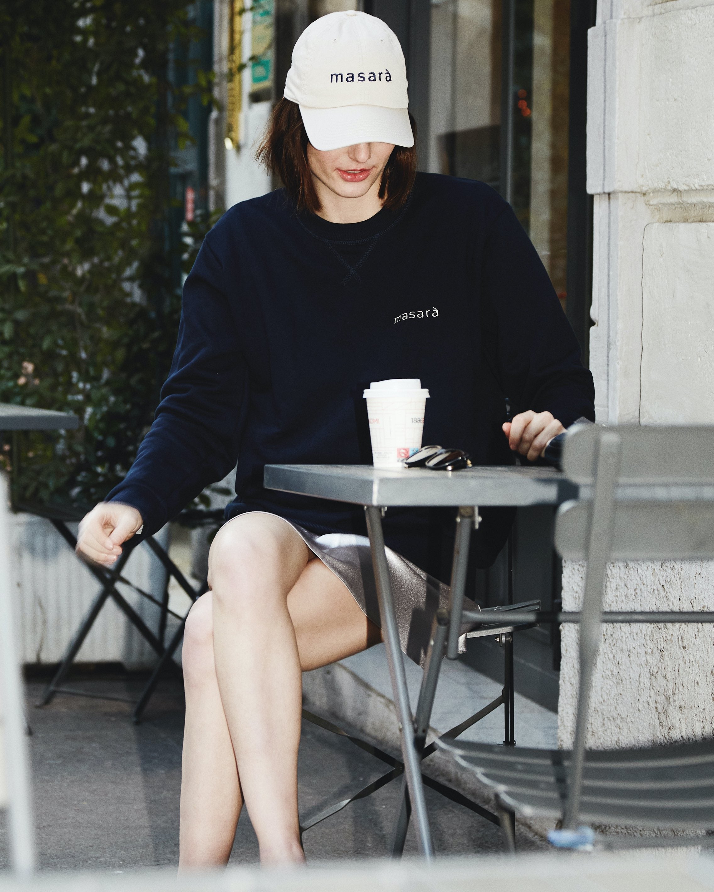 Girl sitting down at cafè table wearing masara beige dad hat and blue sweatshirt