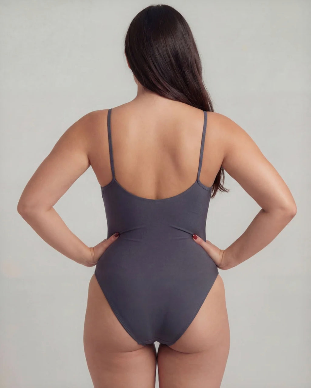 A woman shown from the back wearing a dark gray one-piece swimsuit with thin shoulder straps and a low open back, standing against a light gray studio background.