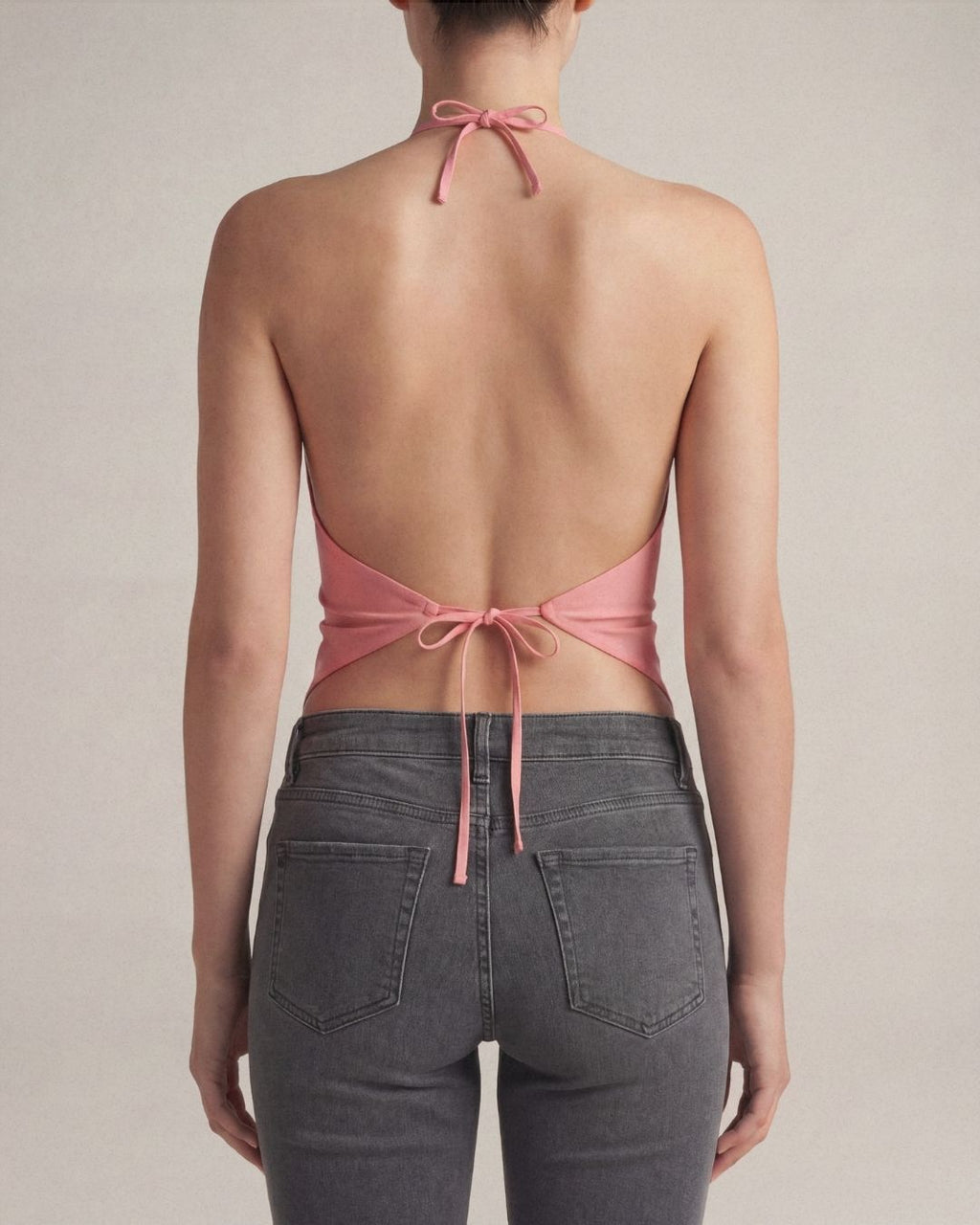 Person wearing a pink halter top with open back and gray jeans on a neutral background