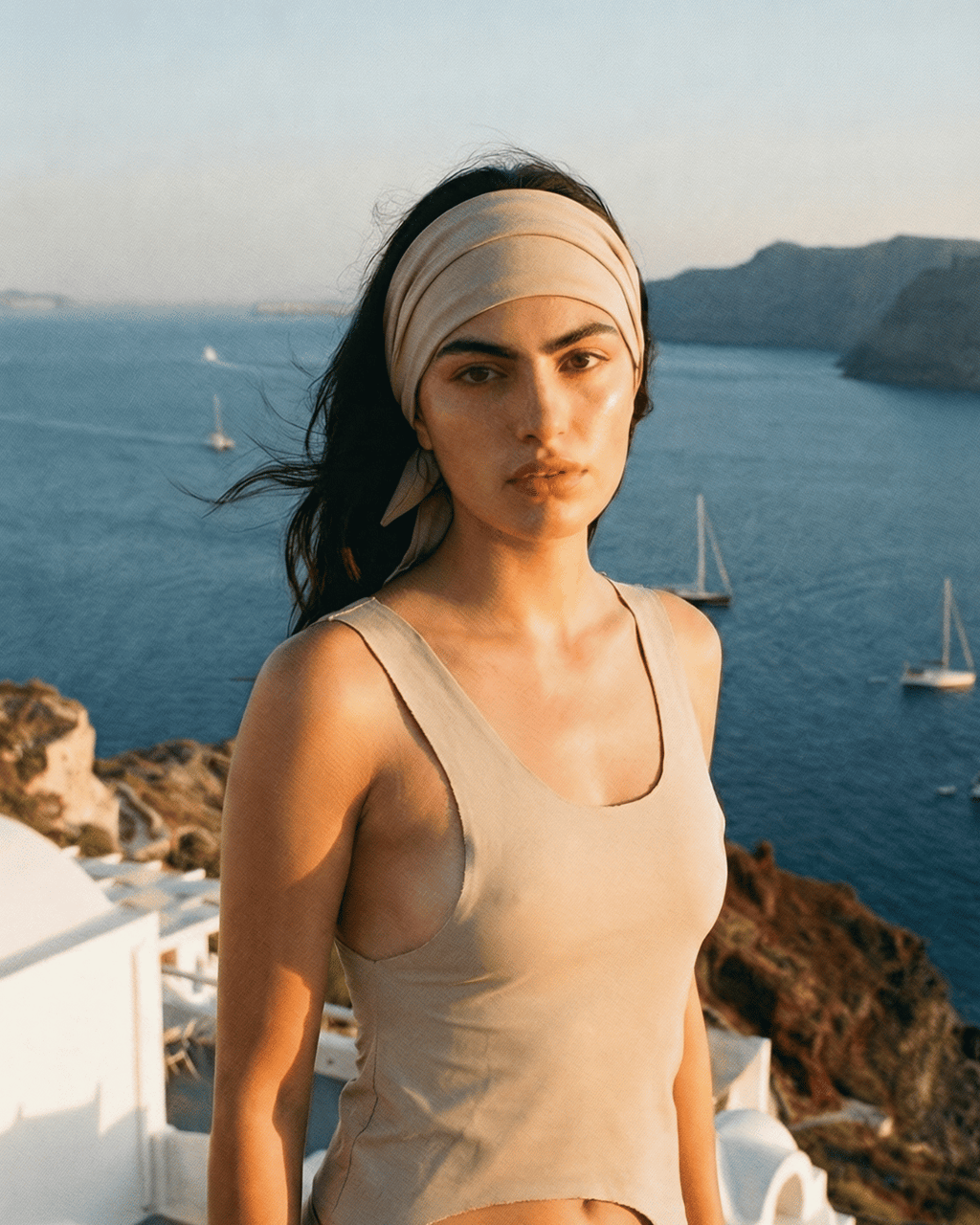 Woman in a beige tank top and headband standing in front of a scenic ocean view with sailboats.