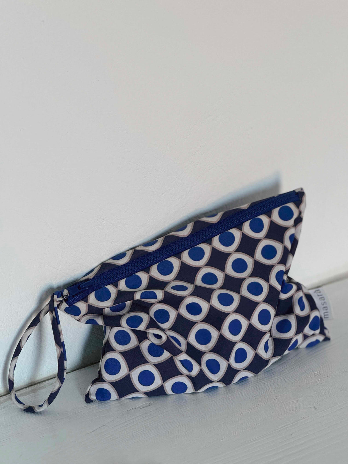 A small zippered pouch with a navy blue, white, and blue geometric pattern, resting on a light-colored wooden surface against a plain white wall.